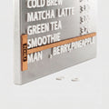 A brushed  Stainless steel  beverage order board with raised white text listing drinks such as cold brew, matcha latte, green tea, smoothie, and berry, pineapple, with a wooden strip holding the time and a small NOKU logo.