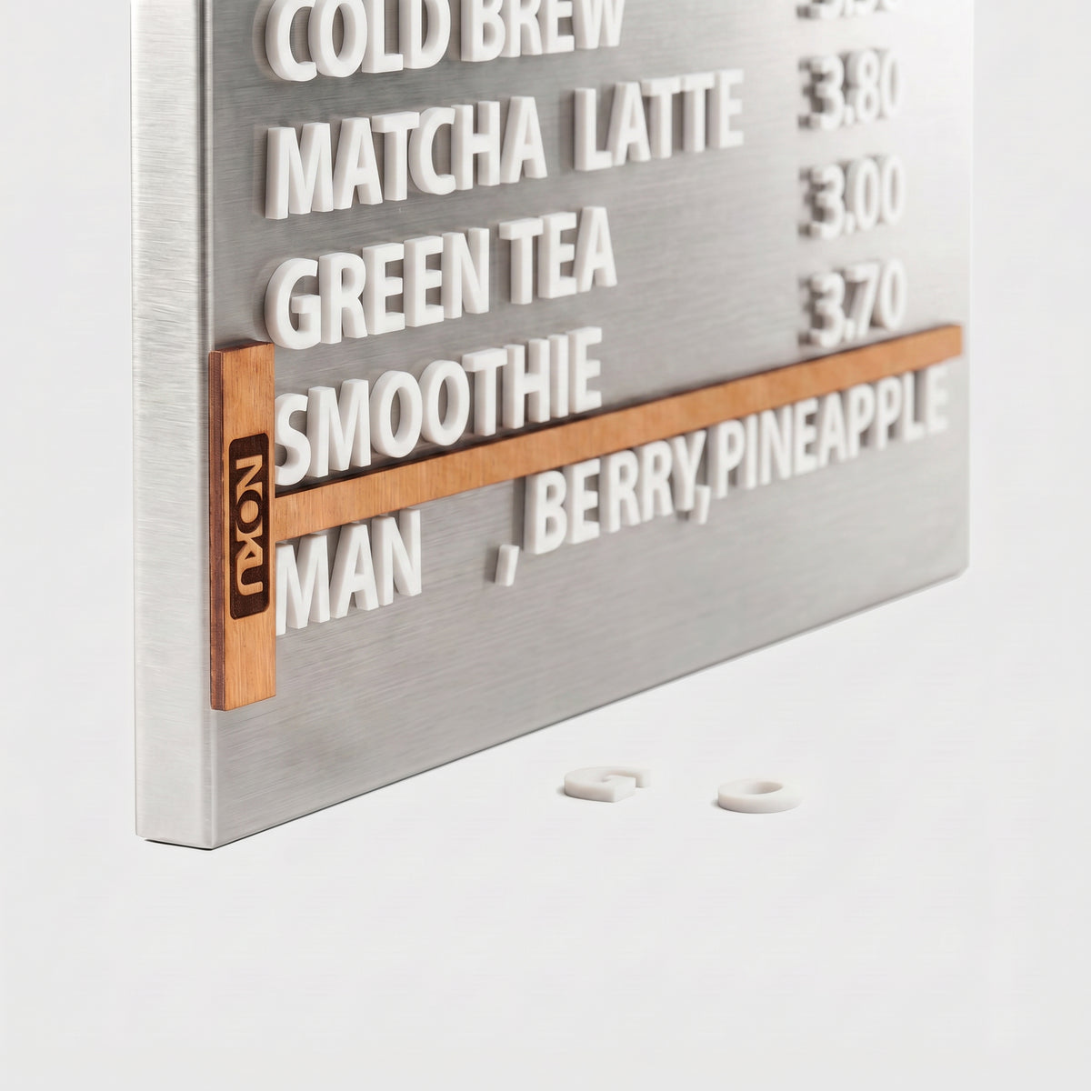 A brushed  Stainless steel  beverage order board with raised white text listing drinks such as cold brew, matcha latte, green tea, smoothie, and berry, pineapple, with a wooden strip holding the time and a small NOKU logo.