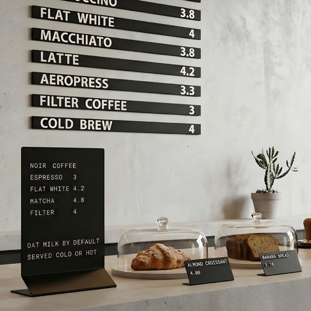 A modern café interior featuring the NOKU display system, which includes a black metal wall menu with white lettering, a tabletop sign with magnetic symbols, and metal price tags with changeable symbols 